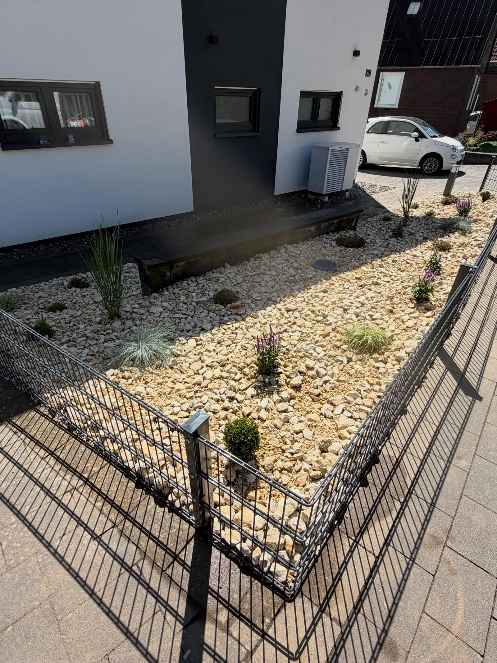 Helles Steinbeet mit kleinen Pflanzen und niedrigem dunklem Zaun vor einer Hausfassade