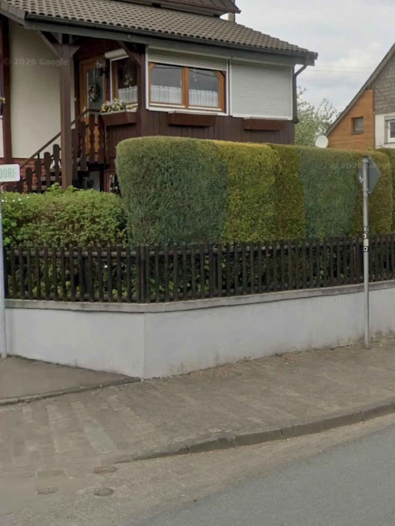 Dunkler Holzzaun auf einer hellen Mauer vor Hecke und Wohnhaus